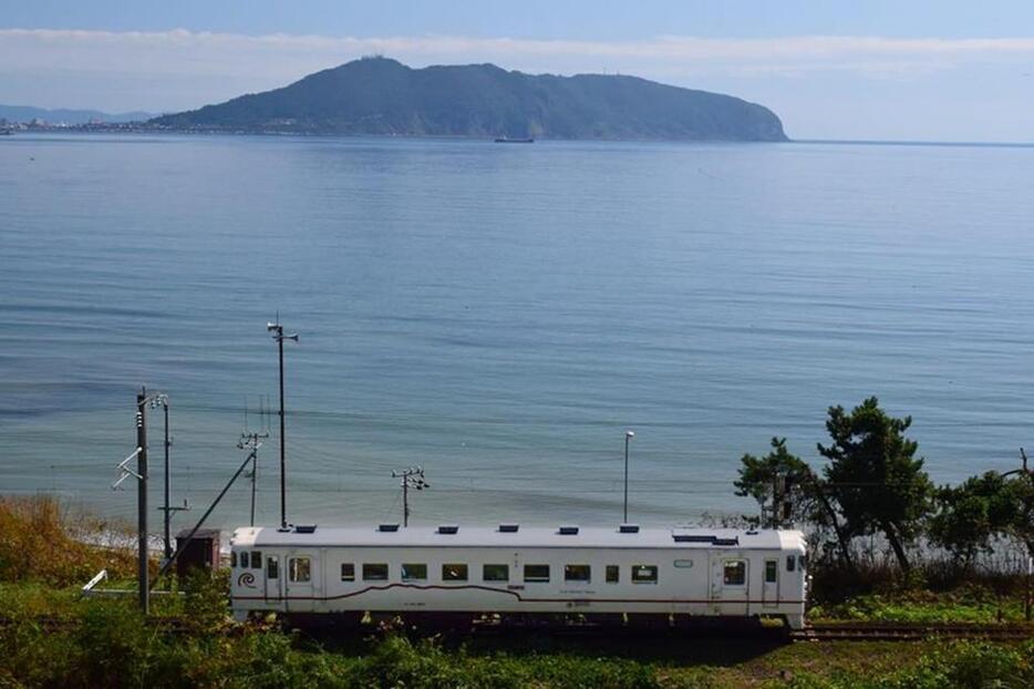 函館山を背景に、函館山の稜線が走る白色の車両　(c)道南いさりび鉄道