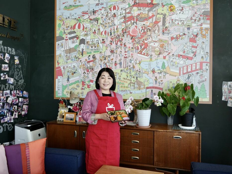 看板商品の「おかん弁当」を手にするオーケーズデリカ3代目の杉本香織さん。背景のイラストがビジョンマップです