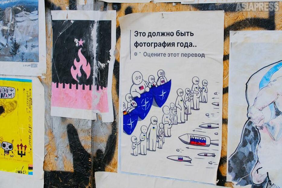 オデーサの街角のポスターが並ぶ壁。この絵の前で足を止める人が少なくない。ロシアのミサイルにさらされるウクライナ市民の後ろでNATOの盾に守られる加盟国。ウクライナのアーチストyeva.hartのイラスト（2024年4月・オデーサ：撮影・玉本英子）