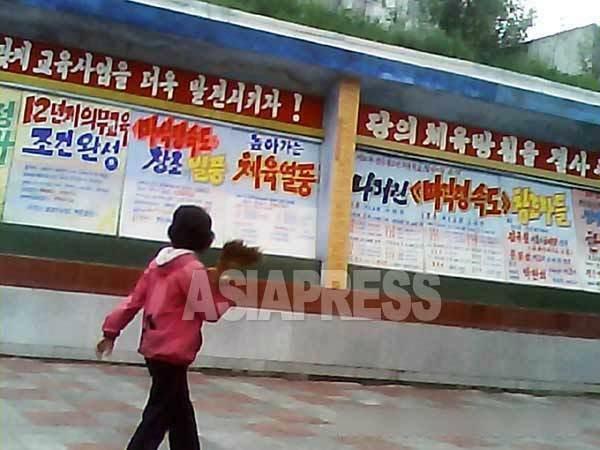 通りの掲示板に掲げられた金正恩政権の12年制義務教育を宣伝する文句。「教育事業をさらに発展させよう！」 というスローガンも見える。2013年8月、北朝鮮北部都市（アジアプレス）