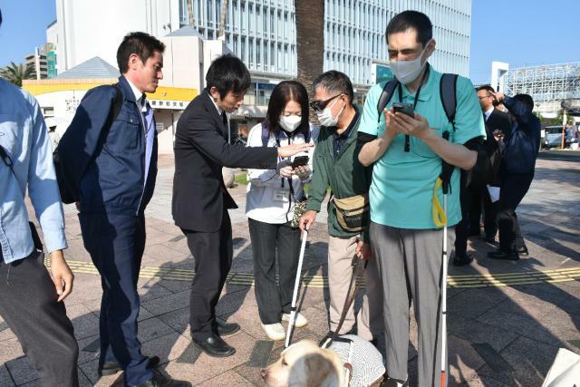 製造メーカー担当者らから、横断歩道歩行支援アプリの説明を受ける視覚障害者ら＝１３日午後、宮崎市・宮崎駅前交差点付近