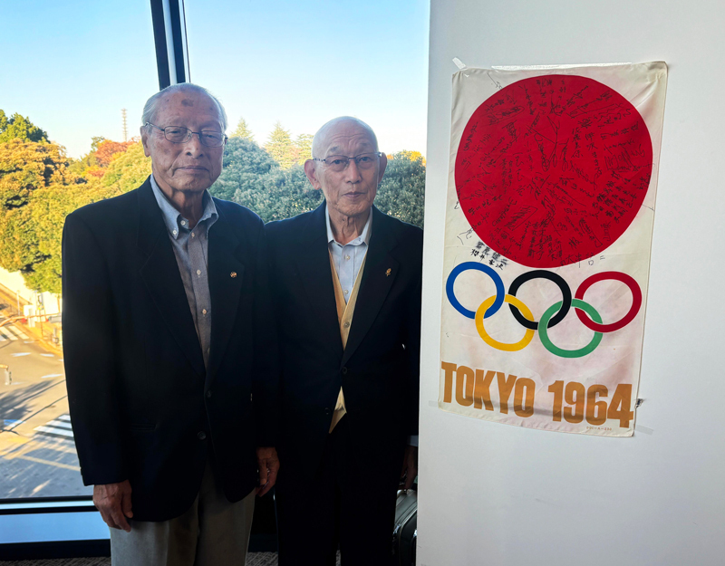 1964年東京五輪陸上日本代表のサインが記された記念のフラッグと写真に収まる五輪3大会連続出場のレジェンド、三段跳の櫻井孝次さん（左）とマラソンの君原健二さん