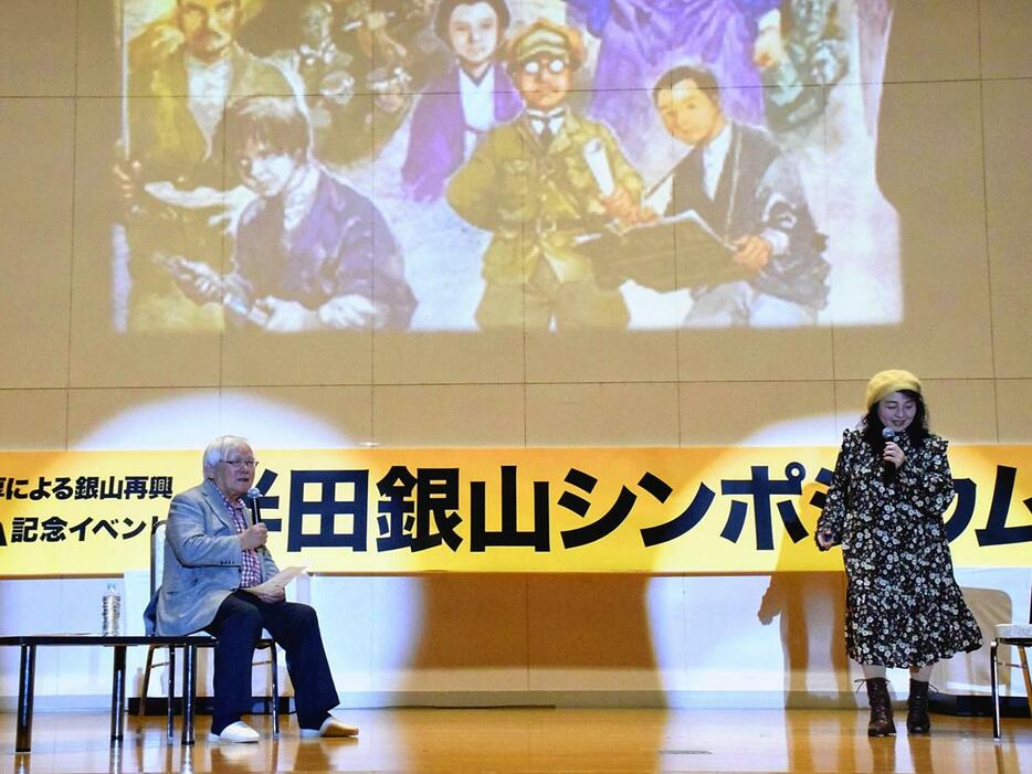 登壇した安彦さん（左）と佐々木さん。後ろのスライドは安彦さんが描いたイベントのポスターの一部。半田銀山で働いていた安彦さんの曽祖父の様子（スライド画像の右端）が描かれている＝10日、桑折町