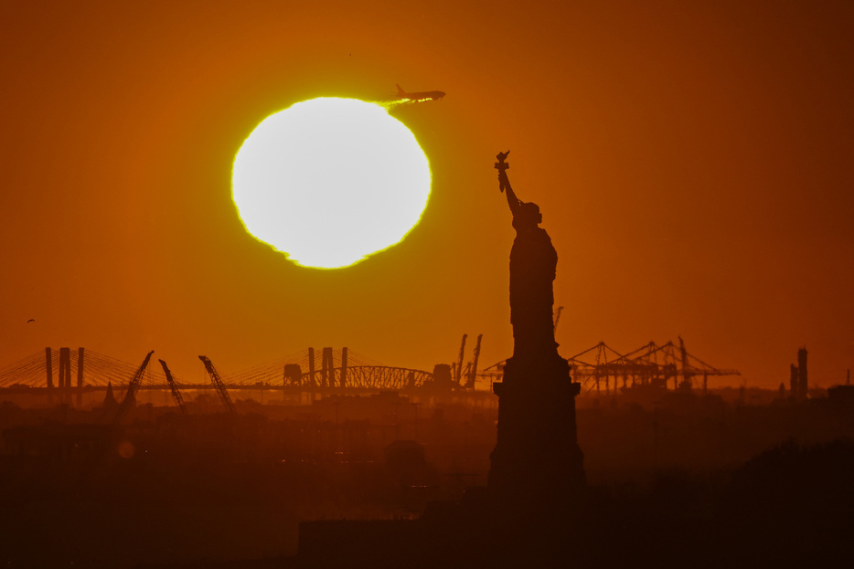 米ニューヨークで、自由の女神像の後ろに沈む夕日に重なる飛行機（2024年11月12日撮影）。【翻訳編集】 AFPBB News