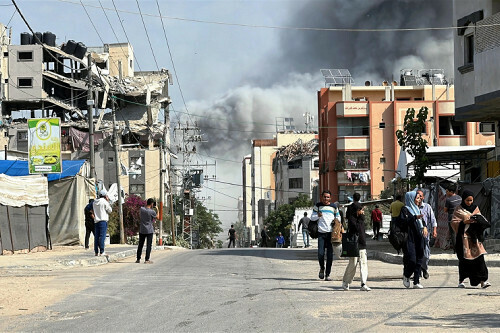 １日、ガザ中部ヌセイラットで、イスラエル軍の攻撃を受け煙が立ち上る中、歩く人々＝ロイター