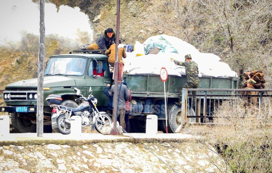 （クリックして拡大）哨所（検問所）の手前で一時停止する木炭車。白い煙が上がっている。エネルギー不足の北朝鮮では、中国からの輸入車を木炭車に改造して使っている。軍隊では冬服に衣替えの時期で、右端の兵士は冬用の軍服を部隊に運ぶものとみられる。