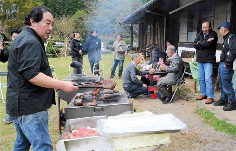 あか牛オーナー制度20周年記念交流会で、肉を調理する宮本健真さん（左）。奥は談笑するオーナーや農家の人たち＝10日、阿蘇市