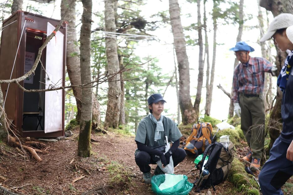 トイレブース（左奥）前で、携帯トイレの使い方を清掃登山の参加者に説明する伊那市職員（左）