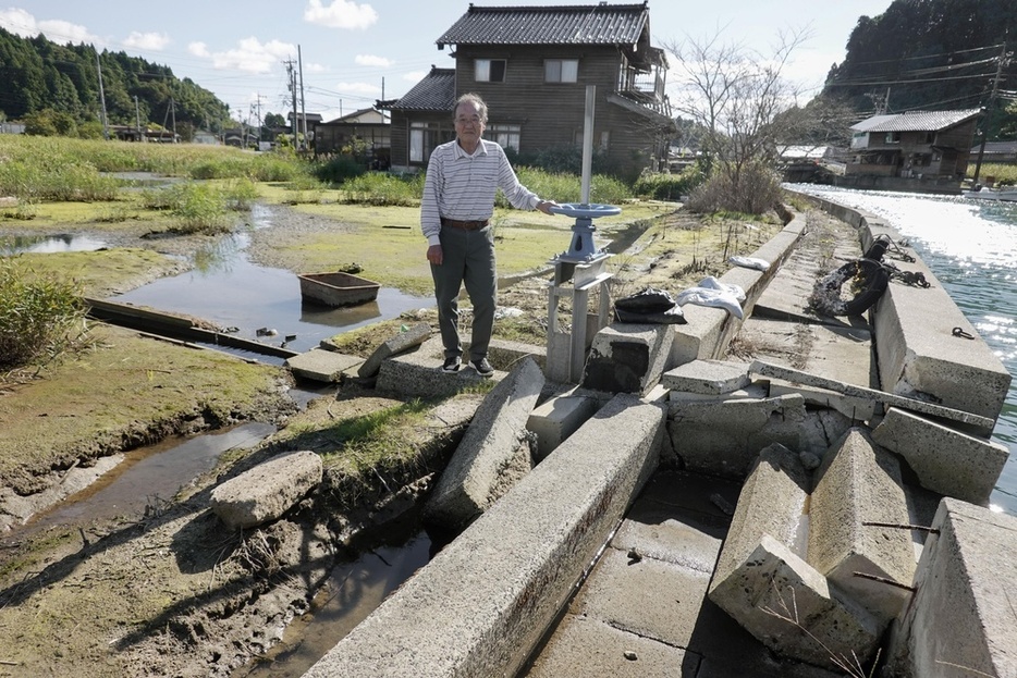 壊れた護岸の脇に立つ泊一夫さん。海水が侵入し水田だった場所は沼と化した。奥は近隣住民の家＝10月13日、石川県穴水町