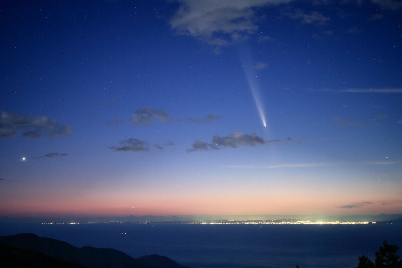 夕暮れの中、長い尾を伸ばす「紫金山・アトラス彗星」。左端は金星、右下は駿河湾を挟んで静岡県焼津市の街明かり＝静岡県西伊豆町で2024年10月14日、手塚耕一郎撮影