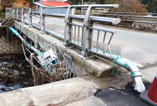 荒瀬川上流の大芦沢川に架かる芦沢橋。橋側面に破損した水道管がぶら下がり、道路脇に仮設管が設置されている＝酒田市北青沢