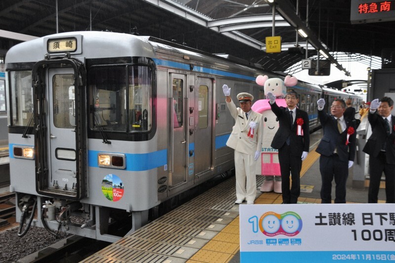 土讃線日下駅～高知駅間の開通100周年を記念するステッカーを付けた列車に出発の合図をする高知市の桑名龍吾市長（中央）ら＝高知市のJR高知駅で2024年11月15日、前川雅俊撮影