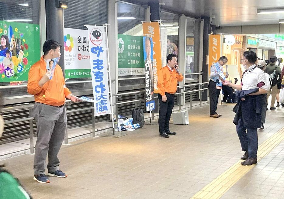 選挙戦初日の夜、山際氏陣営の駅頭活動でマイクを握る秘書とチラシの顔写真を見比べる通勤客。山際氏の姿はなかった＝１０月１５日午後７時２０分ごろ、川崎市中原区