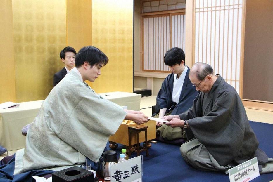 藤井聡太竜王（中）が見守る中、立会人の桐山清澄九段（右）に封じ手を手渡す佐々木勇気八段（左）（日本将棋連盟提供）