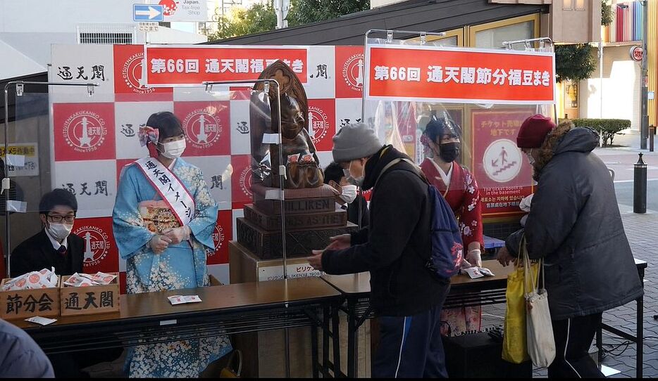 ［写真］通天閣入り口前で行われた福豆配り。ビリケンさんが見守る中、テーブルに福豆を置いて渡す非接触の方式で福豆が配られた。並んでいる人は手の消毒を行っていた＝1日午前10時すぎ、大阪市浪速区で（写真提供：通天閣観光）