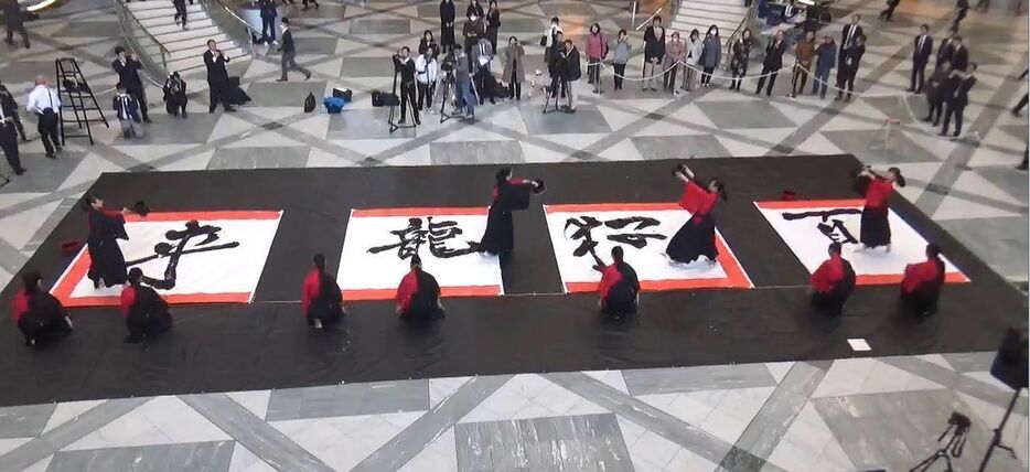 ［写真］多くのサラリーマンや報道陣が見守る中、大きな筆で豪快な踊りを披露する高校生たち＝17日午後0時35分ごろ、大阪市中央区で