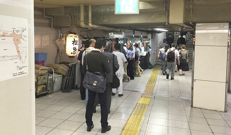 [写真]松葉の行列。左側の囲いは撤去した店を覆うもの。拡幅工事後のイメージ図などが張られていた＝8日午後7時すぎ、大阪市北区で