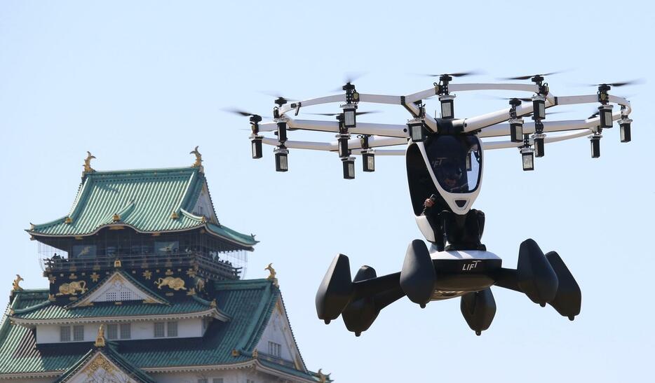 ［写真］大阪城を背にパイロットが乗った状態で飛行する「空飛ぶクルマ」＝14日午後1時40分ごろ、大阪市中央区で