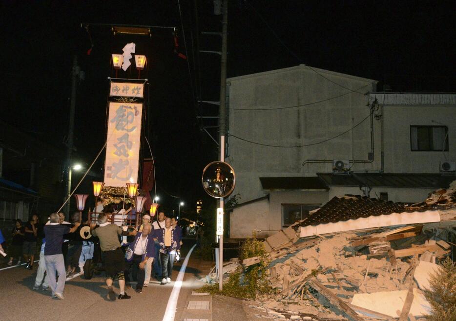 石川県輪島市町野町曽々木地区の夏祭り「曽々木大祭」が開かれ、地震で被害を受けた建物の前を進む巨大な灯籠「キリコ」＝17日夜