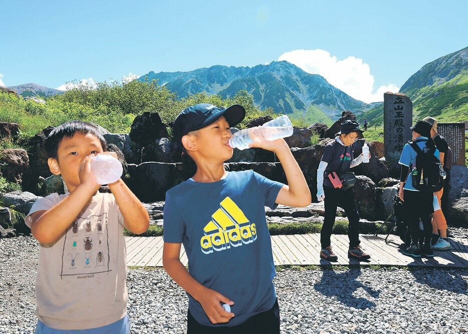 青空の下、立山玉殿の湧水を飲む子どもたち＝立山・室堂