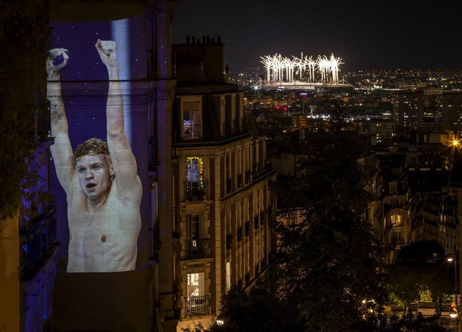 パリ五輪の閉会式が行われたフランス競技場に打ち上がる花火（右上）。左は映し出された競泳フランス代表のレオン・マルシャン＝11日、パリ郊外（ゲッティ＝共同）