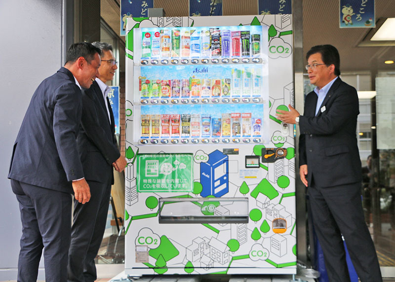 市役所本庁舎の「CO2を食べる自販機」前で歓談する（右から）倉成市長、守谷常務、住吉社長