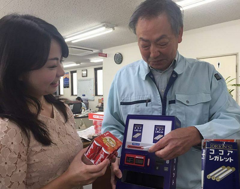 [写真]自動販売機を模したココアシガレットの箱を持つ高岡常務。no smokingと書かれたカードを刺さないと取り出せないという、同社の代表的な菓子でも時代に合わせたパロディを実施=大阪市淀川区のオリオンで