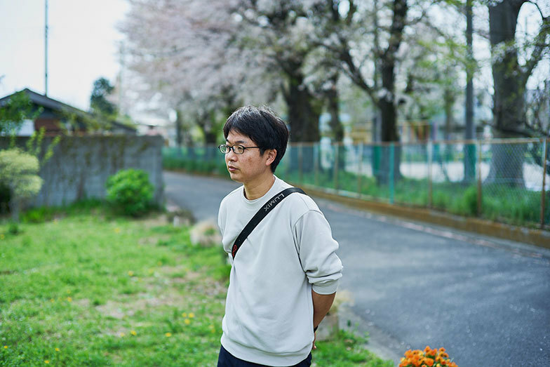 渋谷純平さん。奥の桜が咲いているのが小学校。敷地はプランターでゆるく境界であることを示しています（写真撮影／相馬ミナ）