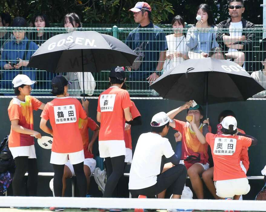 日よけの傘の下で水分補給するソフトテニスの選手＝２５日、鹿児島市の東開庭球場