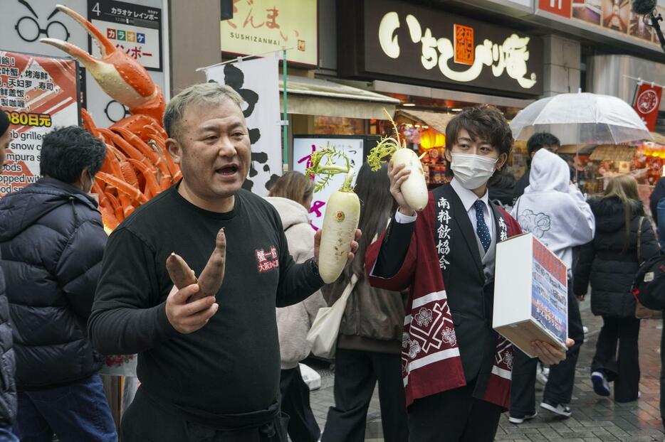 加賀野菜を持ち、能登半島地震の支援を呼びかける武田源社長（左）ら＝23日午後、大阪・ミナミ