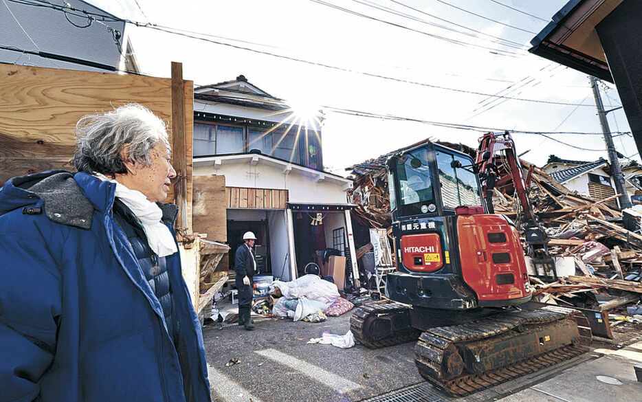 地震で倒壊し、市道にはみ出した自宅の撤去作業を見守る九尾さん＝３１日午後１時半、輪島市河井町