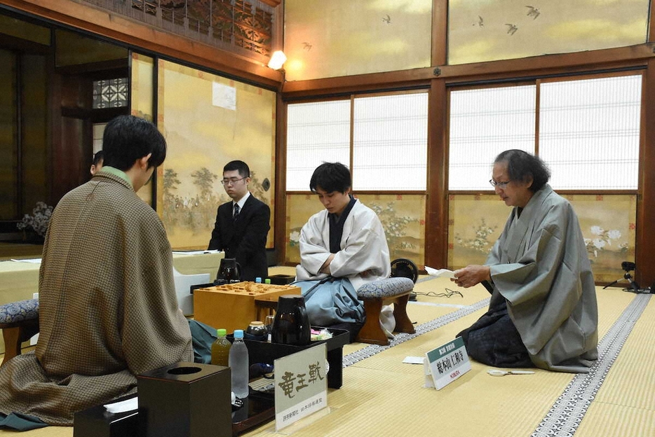 藤井聡太竜王（左）と佐々木勇気八段（右から2人目）へ竜王戦第3局の封じ手を示す立会人の福崎文吾九段（右）（日本将棋連盟提供）