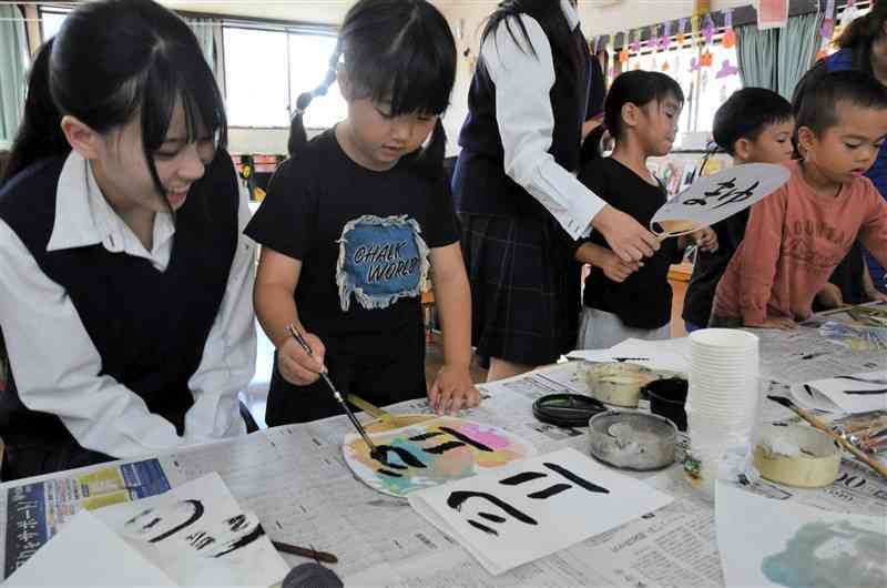 色付けしたうちわに筆で文字を書く園児と、優しく指導する御船高生（左）＝21日、御船町