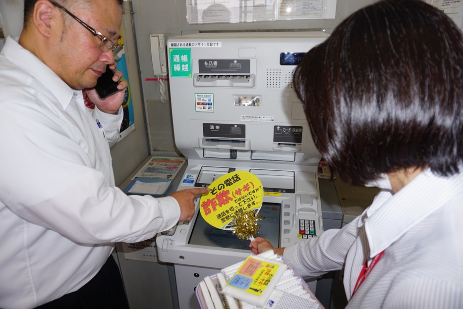 郵便局と篠山警察署の特殊詐欺防止の連携協定を機に始めた、特殊詐欺が疑われるATM前の利用客に局員が啓発うちわを差し出すデモンストレーション＝兵庫県丹波篠山市北新町で