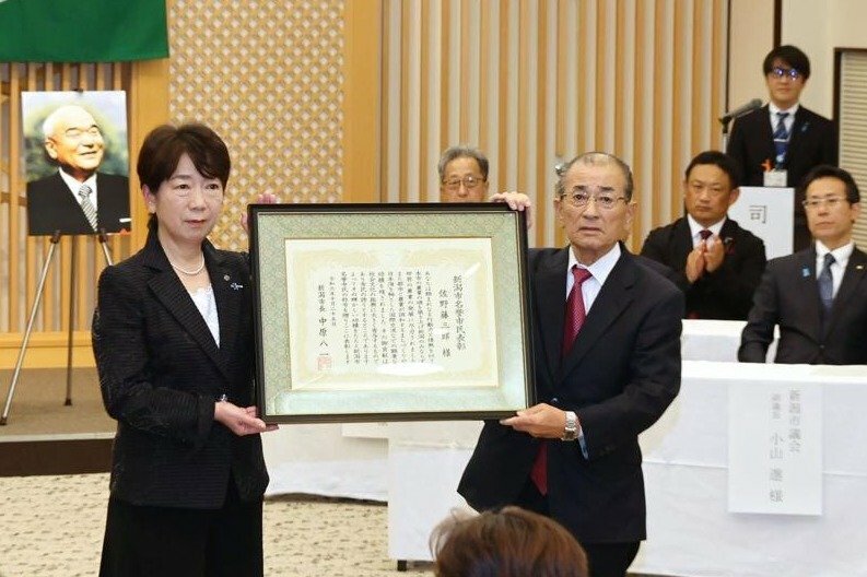 故・佐野藤三郎さんに代わって表彰状を受け取る長男の正人さん（右）＝１０月２５日、新潟市中央区一番堀通町