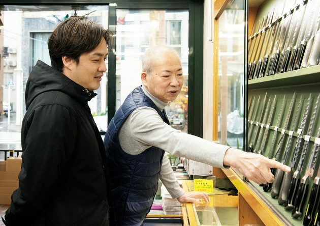 3代目店主で研ぎ師の鎌田さん（右）と山田さん（左）。包丁のデザインを見比べ中。「いい刃物はやっぱりカッコいいんです。」（鎌田さん)「外見や佇まいはとても大事ですよね。」(山田さん)