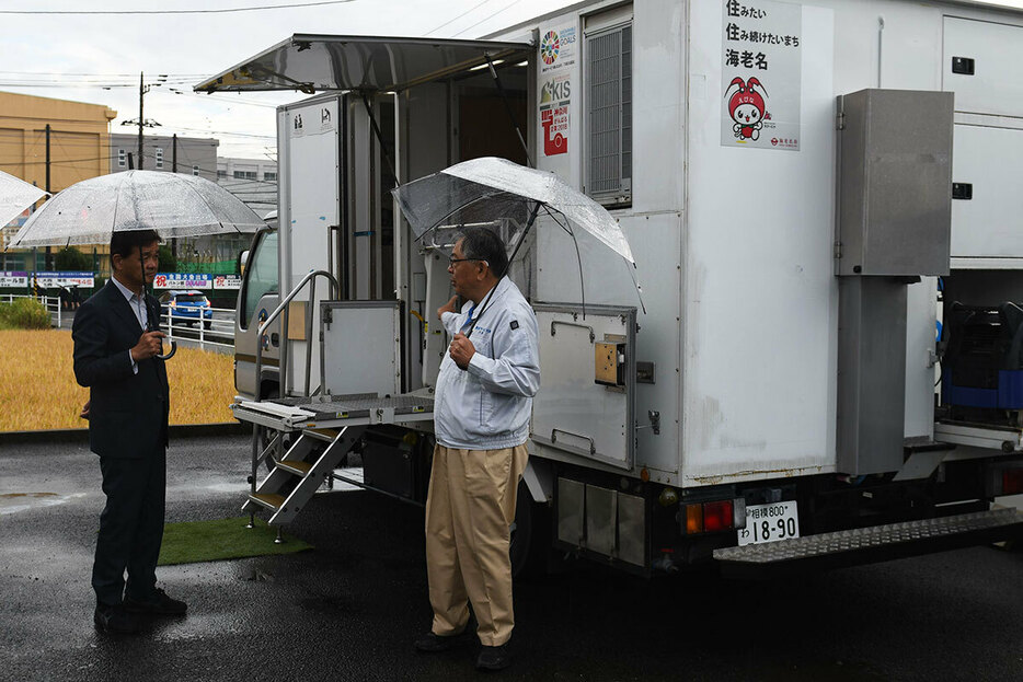 被災地に派遣されてきたトイレカーを前に説明を聞く山神裕町長（左）＝９日、海老名市内