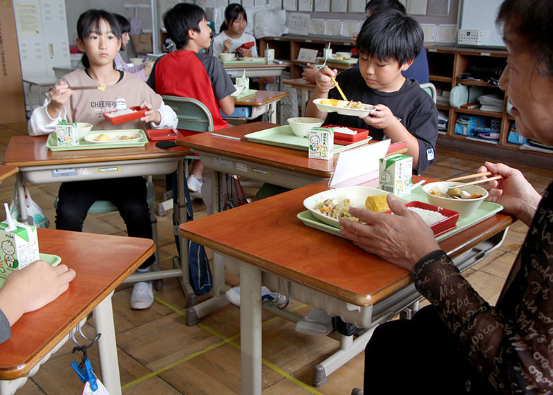 生産者（手前右）の話を聴きながら「町内産食材の日」の給食を味わう児童