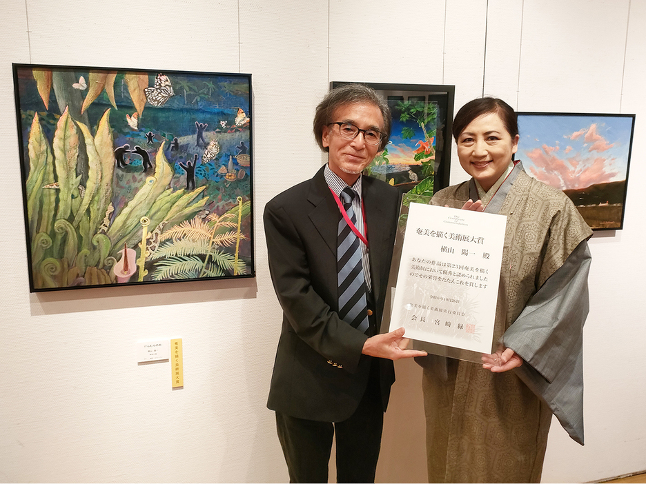 大賞を受賞した横山陽一さん（左）。左の絵が大賞作品「けんむんの社」＝26日、鹿児島県奄美市笠利町