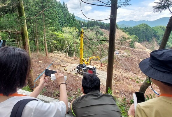 7月17日、高知県仁淀川の山岳地帯である坪井山の頂上付近の伐採作業現場を視察団が見学している。1機当たり9000万円以上する欧州製タワーヤーダが配置されて別の伐木機械が伐採した原木を一列に並べて運搬している