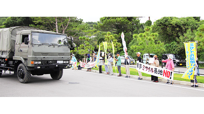 陸自隊員を乗せた車両が駐屯地を出る中、抗議を行う参加者（写真右）＝23日午後1時すぎ、石垣駐屯地