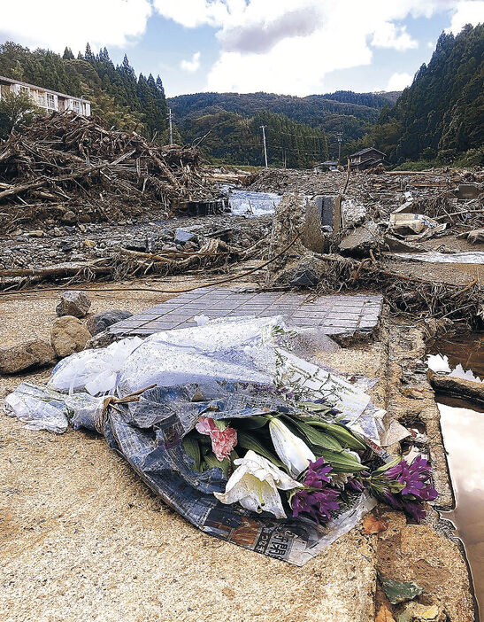 翼音さんが家ごと押し出されたとみられる自宅跡。数日前に供えられた花束は雨でぬれたままだった＝１１日午前１１時半、輪島市久手川町