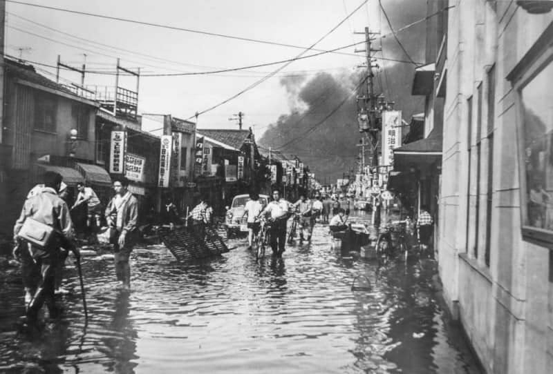 冠水した道路を歩く人たち。石油タンク火災の黒煙も見える＝１９６４年６月、新潟市（近藤武夫さん提供）