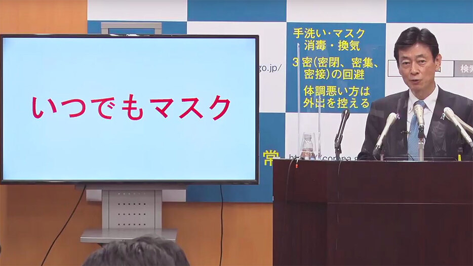 「いつでもマスク」と表示された画面の横で説明する西村担当相