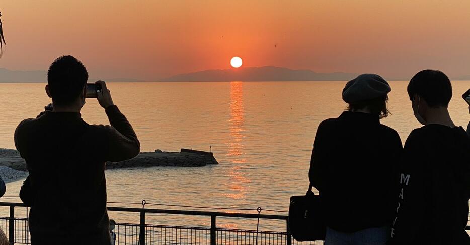 ［写真］雲ひとつないさわやかな天気のもと、まんまるな夕陽を眺める若者たち。「淡路島に沈む感じがいい」と話しながら写真撮影を楽しんでいた＝12日午後4時45分ごろ、大阪府泉佐野市で