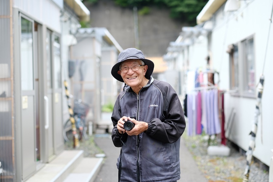 住み慣れた仮設住宅の前で、カメラを手にする小川さん（撮影：山本宏樹/deltaphoto）