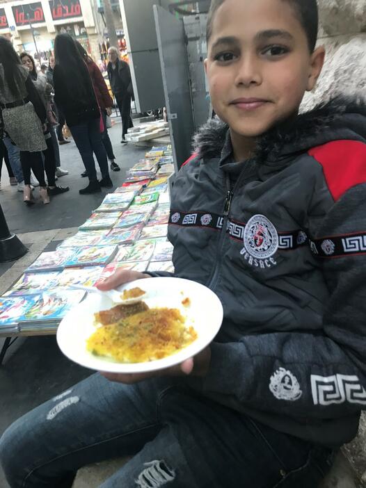 さくさくしたペイストリー生地の上に伸びの良いチーズがのった人気のアラブ菓子「カナーフェ」を持つヨルダン人の少年。アンマンの街中で出会った人々の多くが、気さくな笑顔を向けてくれた。2019年12月19日。（撮影・村松まどか）