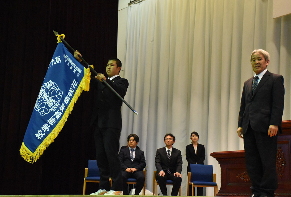 田中一夫校長（右端）から受け取ったセンバツ旗を掲げる井上朋也主将＝花咲徳栄高校第１体育館で