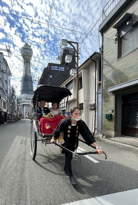 ［写真］通天閣を背に男性2人を乗せて人力車で駆ける板倉さん＝大阪市浪速区で