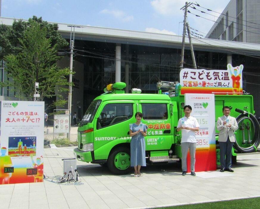 えきまえテラスに設置される「こども気温」を知らせる緑色の消防車「熱中症防車」。右から八王子市の初宿和夫（しやけ かずお）市長、サントリー食品インターナショナルの阿部泰丈ブランドマーケティング本部部長、多胡氏